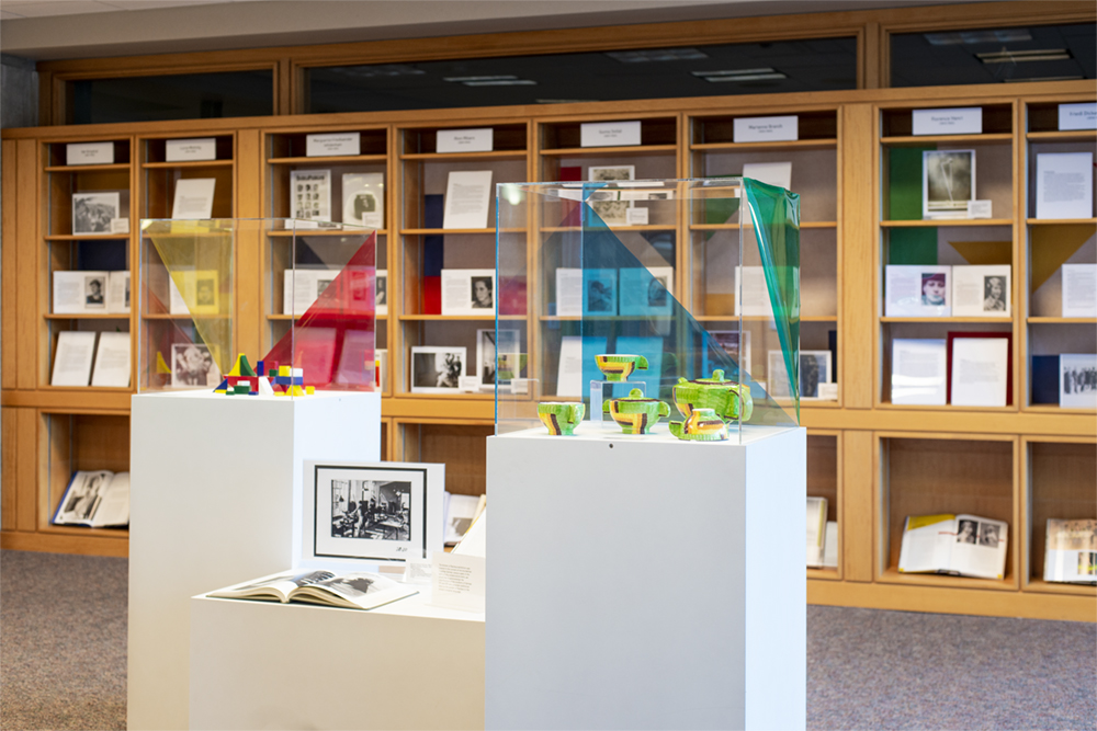 Shelf in the background with students work that tell stories about the women of Bauhaus in german. In front are a series of pedestals that showcase art work and pieces that were created for the show as well as curated. Books on the pedestal tell the story of women in Bauhaus.