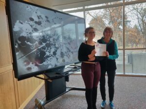 Maddison Jones with Judge Accepting her first place award for the art competition in front of a TV displaying her work - a black and white abstract photograph with circles and lines and reflections.