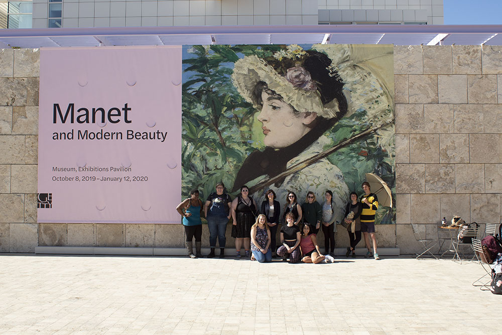 Large billboard poster with the title Manet and Modern Beauty. Image of a woman wearing a bonnet and holding an umbrella. In front of the billboard is a group of students posing for a photo.