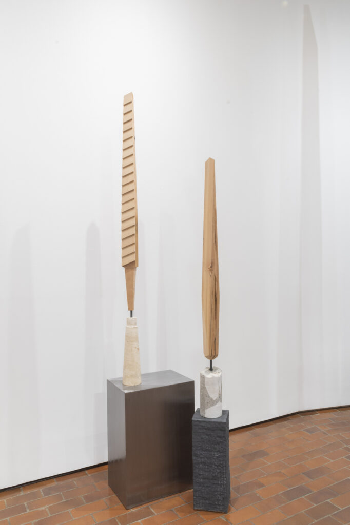 Two tall carved wooden sculptures on white pedestals in a white gallery corner.