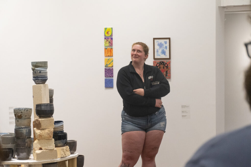 A man dressed in black stands with his arms crossed in front of a white gallery wall with framed artworks.