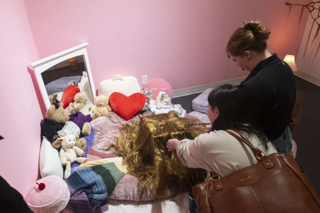 A person with long, light-brown hair leans over a collection of stuffed animals and soft toys arranged against a pink wall, with a red heart-shaped cushion among them.