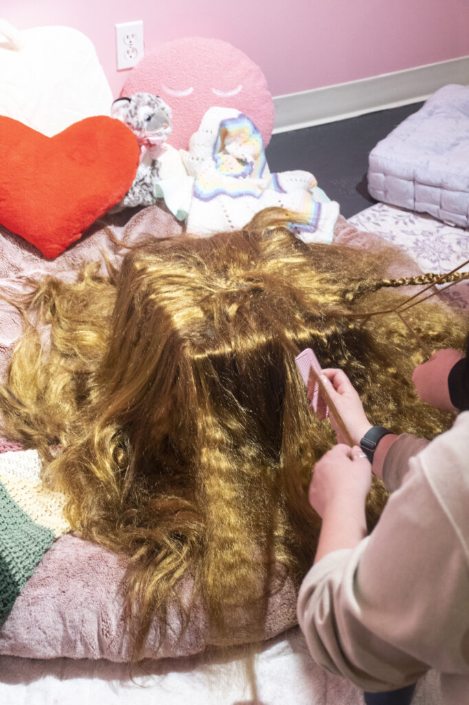 A person with long, light-brown hair leans over a crowded display of stuffed animals and soft toys, including a prominent red heart-shaped cushion.