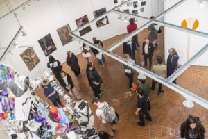 An overhead view of a group of people gathered in an art gallery, with framed artworks displayed along the walls and visitors standing or sitting around the exhibition space.