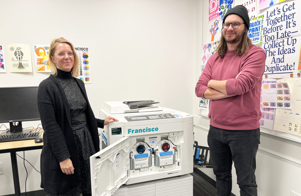 Two people standing in front of the riso machine.