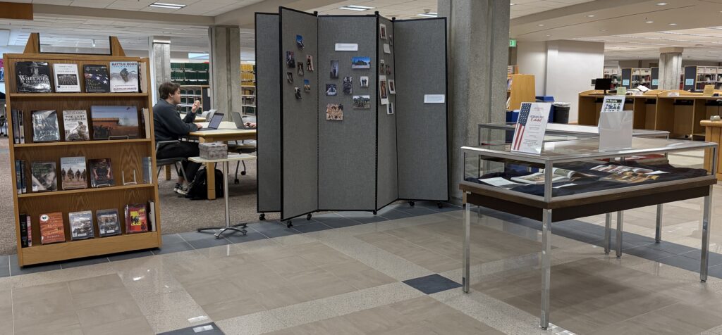 photo of the Veterans Day display in the lobby of JFK Library. Display includes recent books, a wall to offer remembrances, and two display cases of graphic novels from the Global War on Terror era.