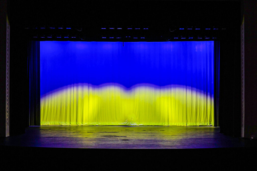 Image of Showalter Auditorium stage lighting arranging to shine the Ukrainian flag with blue and yellow reflecting on the stage curtain