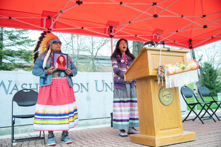 Photo of tribal leaders dressed in traditional clothing. One is holding up a picture of her sister who died under suspicious circumstances.