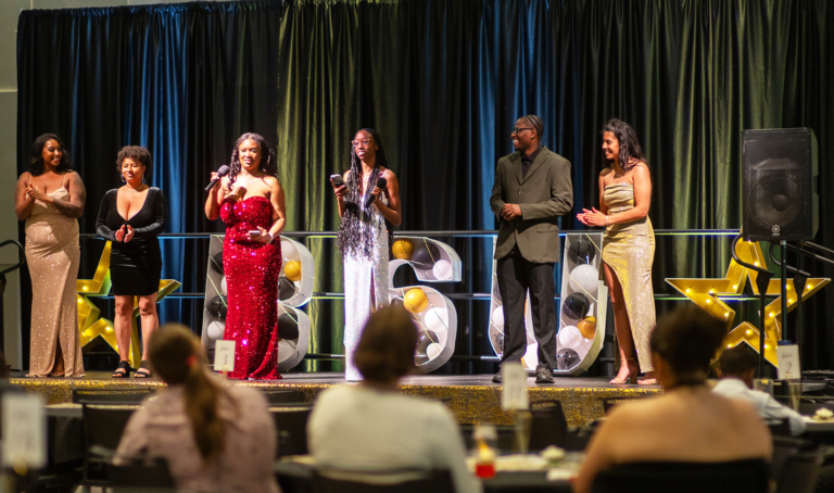 The two emcees on the stage with four guests with everyone wearing dress attire.
