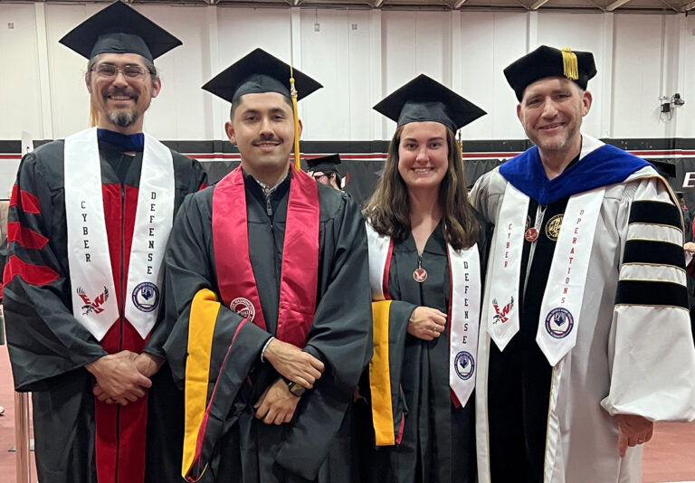 Two cybersecurity students in caps and gowns along with two gowned faculty members.