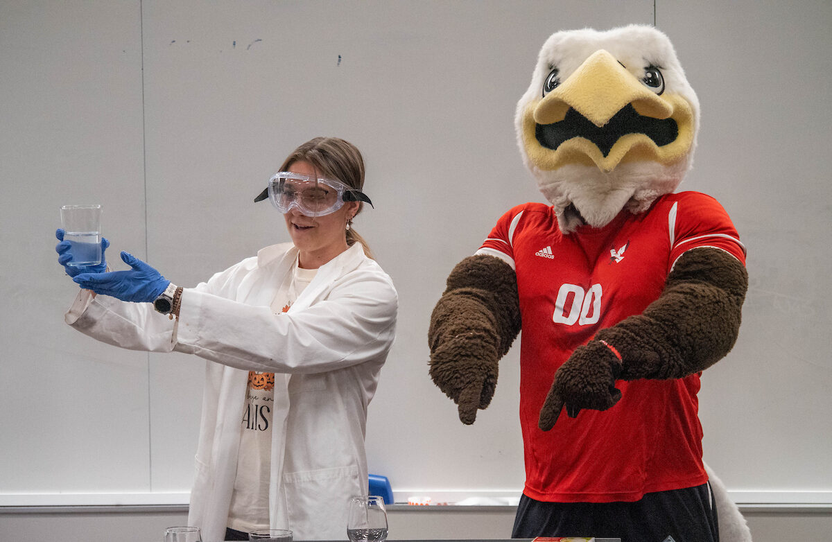 Student in lab coat demonstrating science project with Swoop helping out.