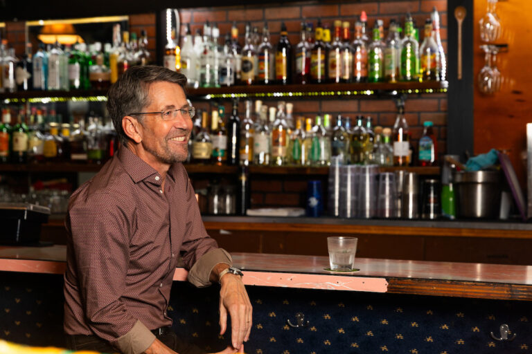 Jess sitting at a counter in an empty bar looking off camera.
