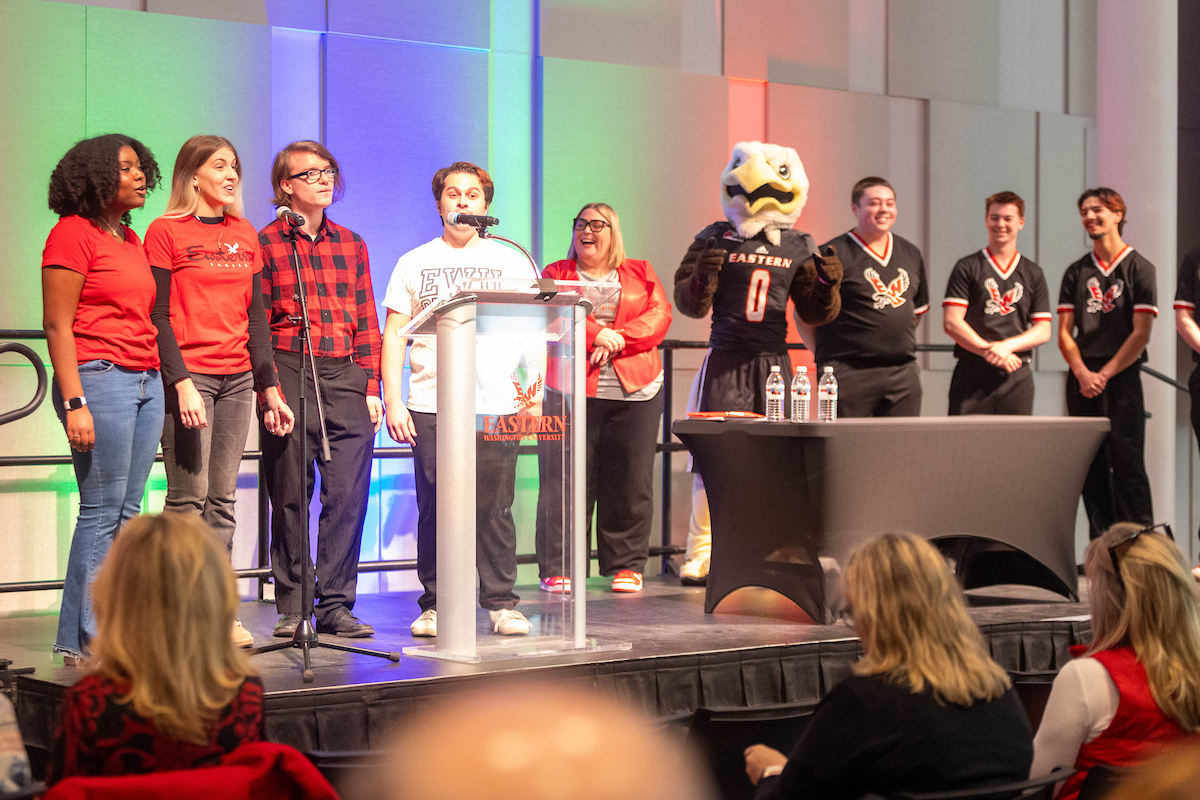 Four person student choir sings on stage with the director of alumni relations, Swoop and three members of the cheer squad nearby.