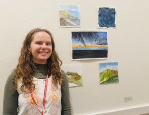 Student standing in front of five watercolor paintings hanging on a wall.