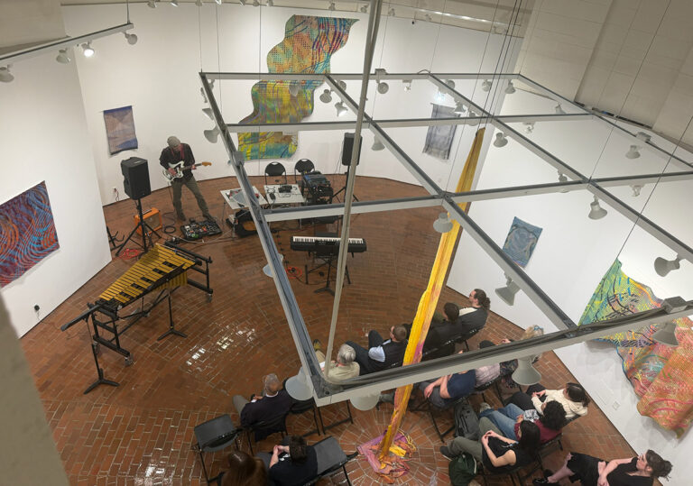 Shot looking down at Art Gallery with musician playing guitar and attendees in seats watching.