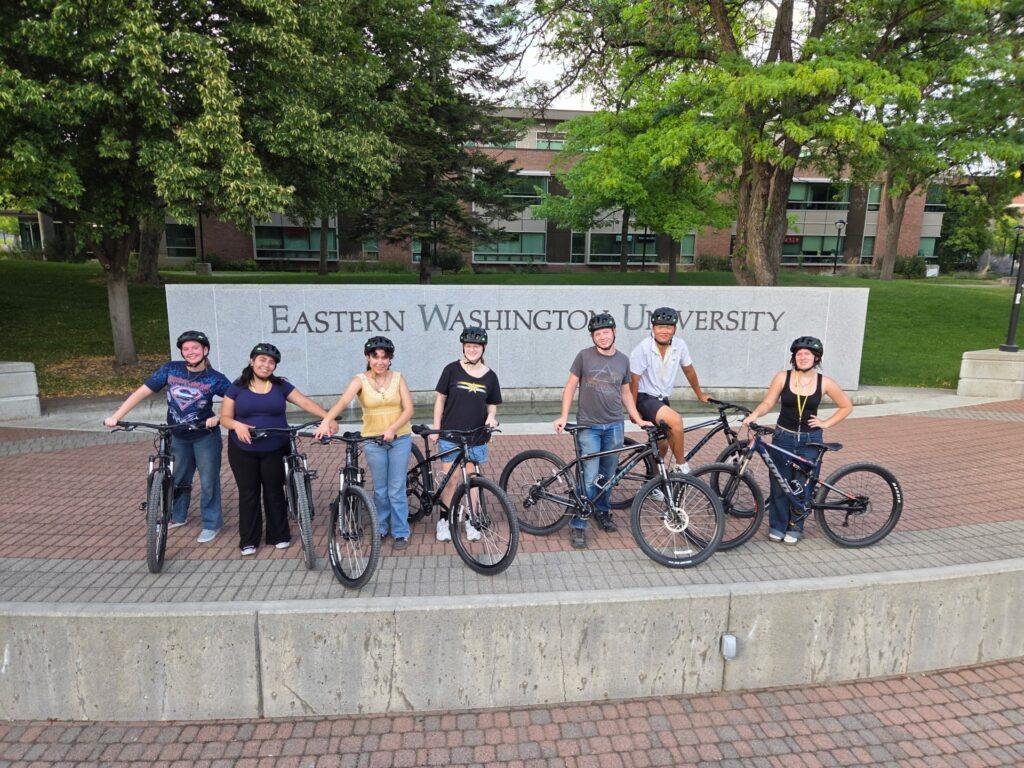 Seven people posing with their bikes in front of the large EWU sign.